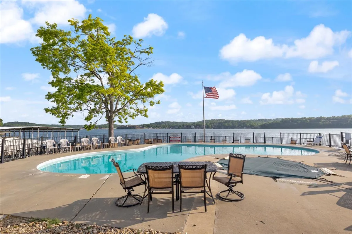 A sparkling swimming pool overlooks the lake, framed by patio furniture, a green tree, and the American flag. The lakeside setting is inviting, with lounge chairs ready for sunbathing or relaxing and the water shimmering in the background, embodying the resort-style amenities described.