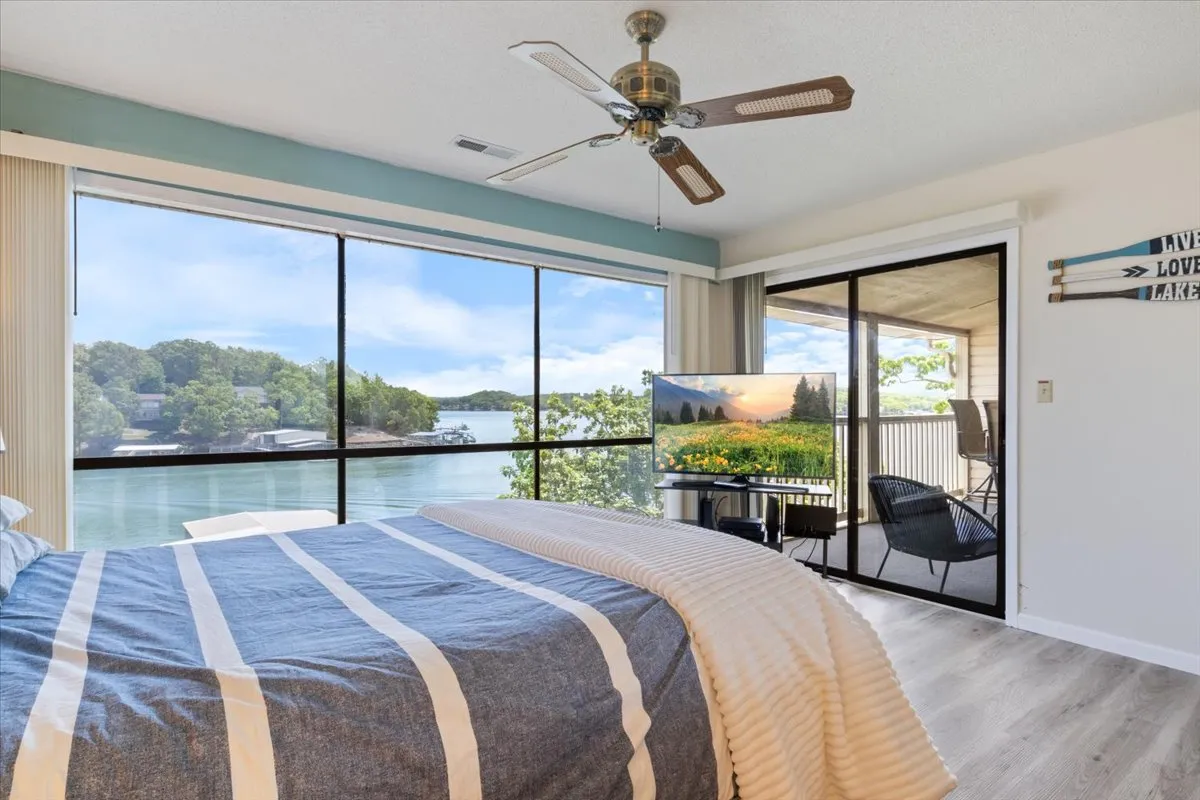A serene bedroom featuring a striped bedspread, large windows offering stunning lake views, a TV displaying a scenic nature scene, and a comfortable seating area on the balcony. The space feels cozy and inviting, ideal for both couples and families.