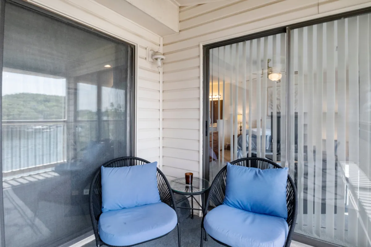This exterior image features two blue cushioned chairs and a small round table on a patio, positioned to overlook the water. Sliding glass doors connect the outdoor seating area to the condo interior, and the tranquil setting suggests an ideal spot for morning coffee, reading, or taking in sunrise views.