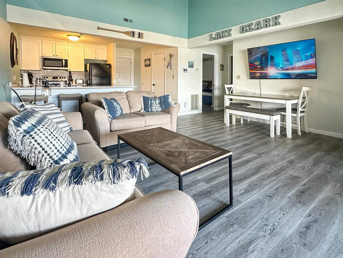 A cozy and inviting living area with a patterned-cushion sofa, wooden coffee table, and a large TV on the wall. The adjacent dining area with white chairs is visible, reinforcing the open layout and sense of togetherness. The space is styled for comfort and socializing.