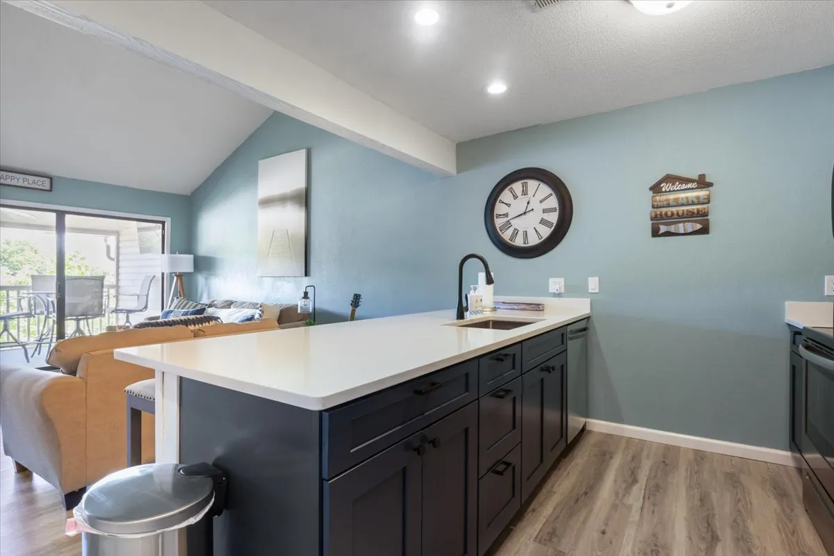 A welcoming kitchen and living area with a kitchen island, clock, welcome sign, cozy sofa, and sunlit windows. The layout allows for both cooking and relaxation, with space for entertaining and daily routines.