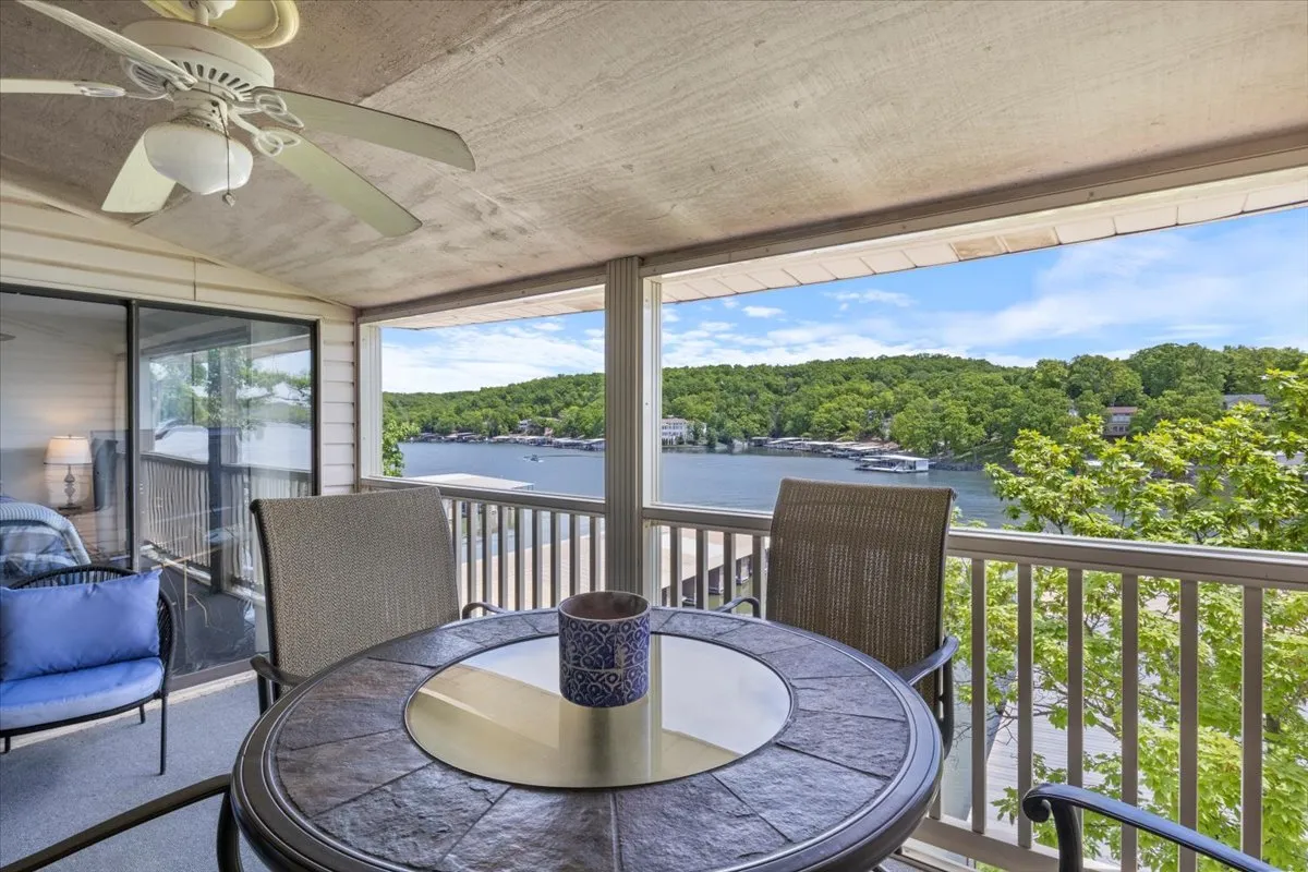 This image highlights an outdoor dining table set on a private deck, surrounded by lush greenery with a tranquil lake view in the background. A ceiling fan provides comfort, while the seating invites guests to dine or relax outdoors. The scene captures the serene atmosphere and the connection between indoor comfort and outdoor living.