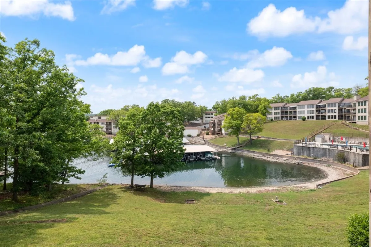 A wide exterior shot showing the lake, a boathouse, surrounding trees, and residential buildings nearby. The lake is calm, and the early morning or dusk lighting adds a peaceful vibe. The scene also hints at activities like fishing, boating, and lakeside picnics, as families might gather on the docks or grassy areas.