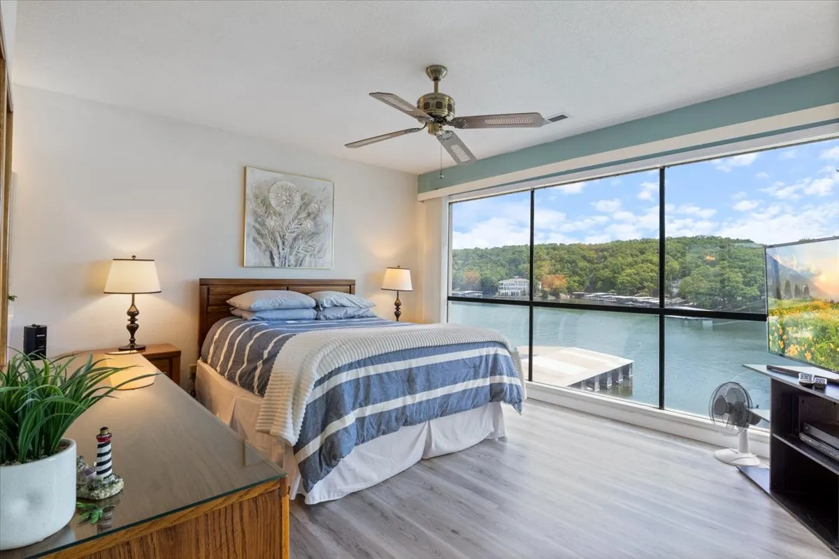 A calming bedroom with a plush bed, large windows overlooking the water, a nightstand with a lamp, and tasteful decorative wall art. The serene atmosphere is enhanced by soft, natural lighting and view of the lake, inviting restful sleep and quiet reflection.