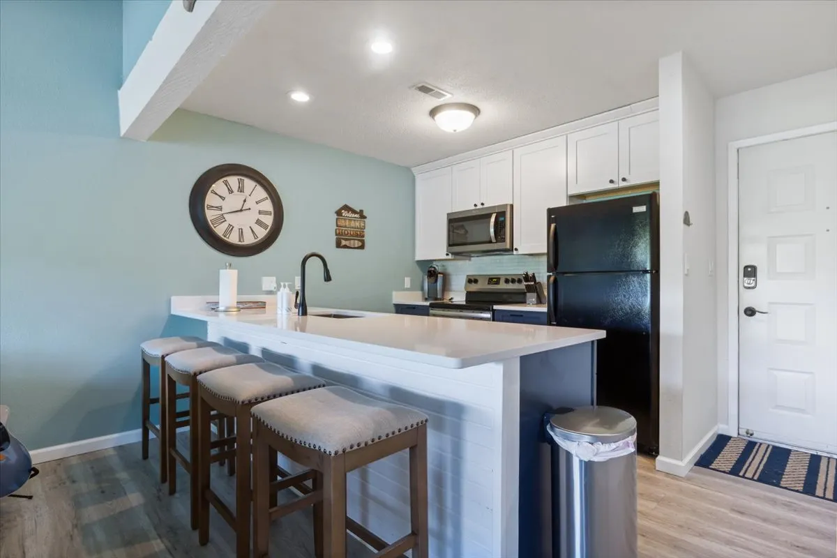 A welcoming kitchen scene with a prominent kitchen island, bar stools, wall clock, and modern appliances. The space is designed for entertaining and casual dining, echoing the functionality and style described in the section and referencing the Pack Less Program.
