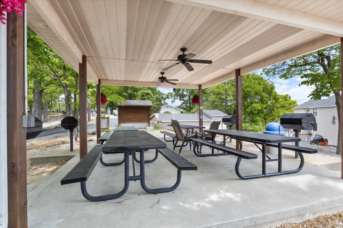 A shaded picnic area features large picnic tables, ceiling fans, hanging flower baskets, and a grill area. The inviting space is perfectly suited for outdoor meals, family gatherings, and leisurely barbecues, offering both shade and a welcoming social atmosphere.
