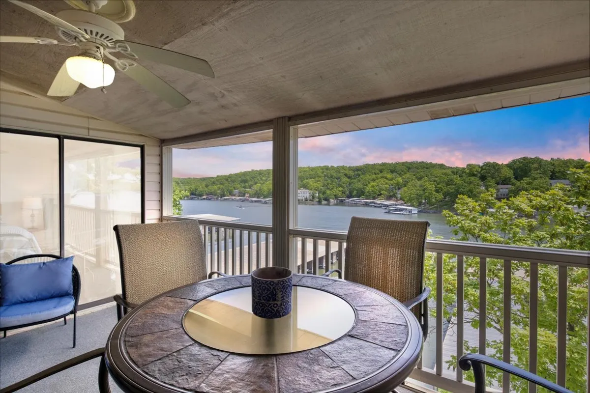 The image features an inviting outdoor dining table set with a ceramic cup, positioned on a deck overlooking the sparkling waters of Lake Ozark. Lush greenery frames the view, creating a tranquil setting perfect for morning coffee, relaxing with a book, or gathering with friends and family as the sun sets over the lake.
