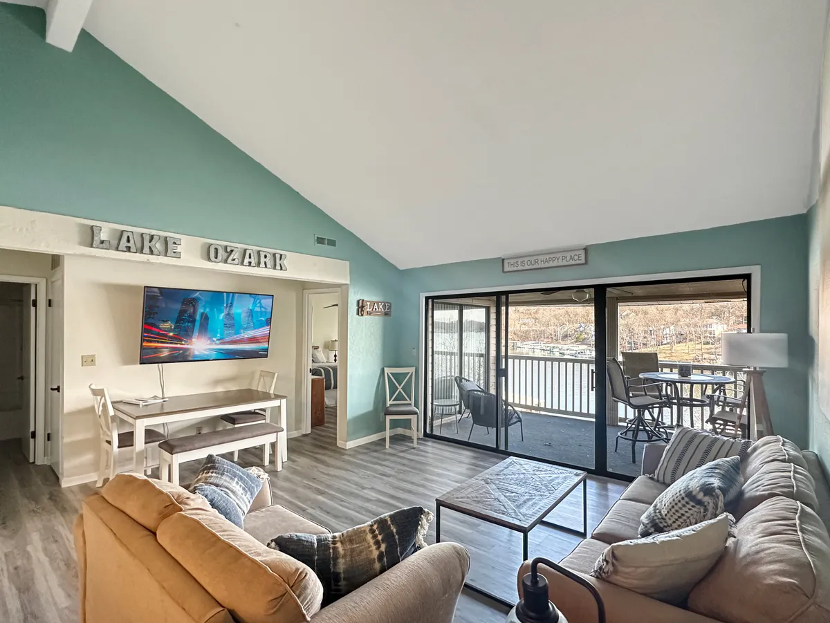 A cheerful and relaxed open-concept living area featuring large windows overlooking the lake. The room is furnished with a comfortable seating area, a TV mounted on the wall, and decorative signage, all bathed in natural light that highlights the tranquil blues and expansive views.